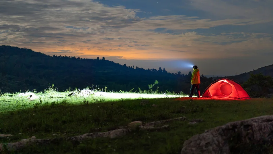 Person with headlamp by illuminated red tent, night camping in mountains under twilight sky.