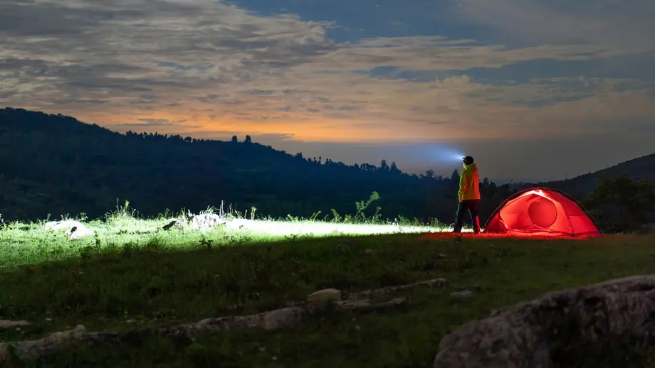 Person with headlamp by illuminated red tent, night camping in mountains under twilight sky.