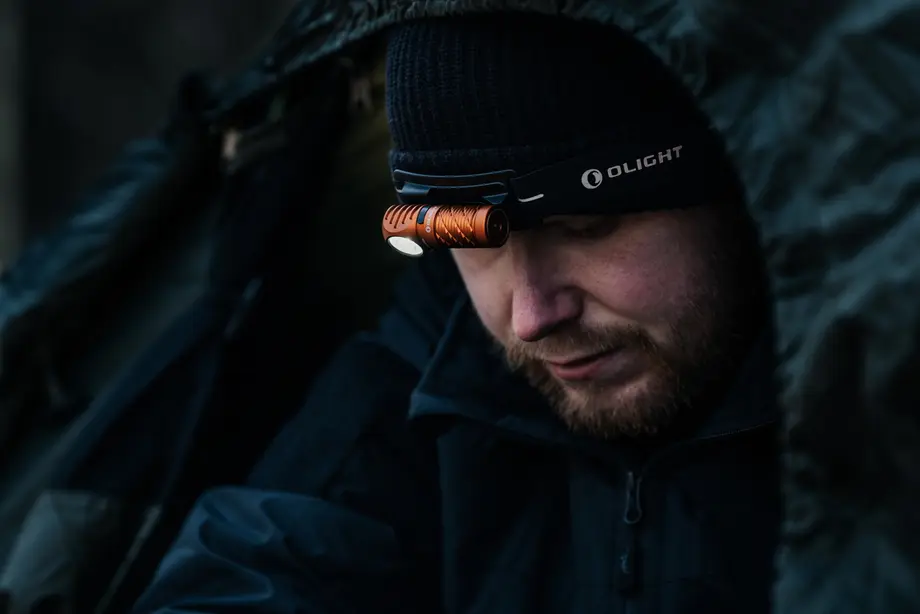 Bearded man wearing black beanie with illuminated orange Olight headlamp, looking down.