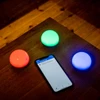 Three smart dome lights glowing orange green and blue next to a smartphone displaying an authenticator app on a wooden table