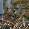 Person standing beside a bike with a saddle bag, red frame and knobby tires in dry grass.