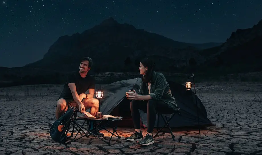 Starry night camping scene: couple sitting by tent with lanterns on cracked earth, mountains in background.