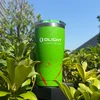 Green Olight insulated travel tumbler with red splatter pattern outdoors in green bushes under blue sky