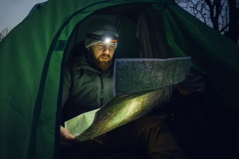 Bearded man in green tent using headlamp to read a topographic map for night navigation.