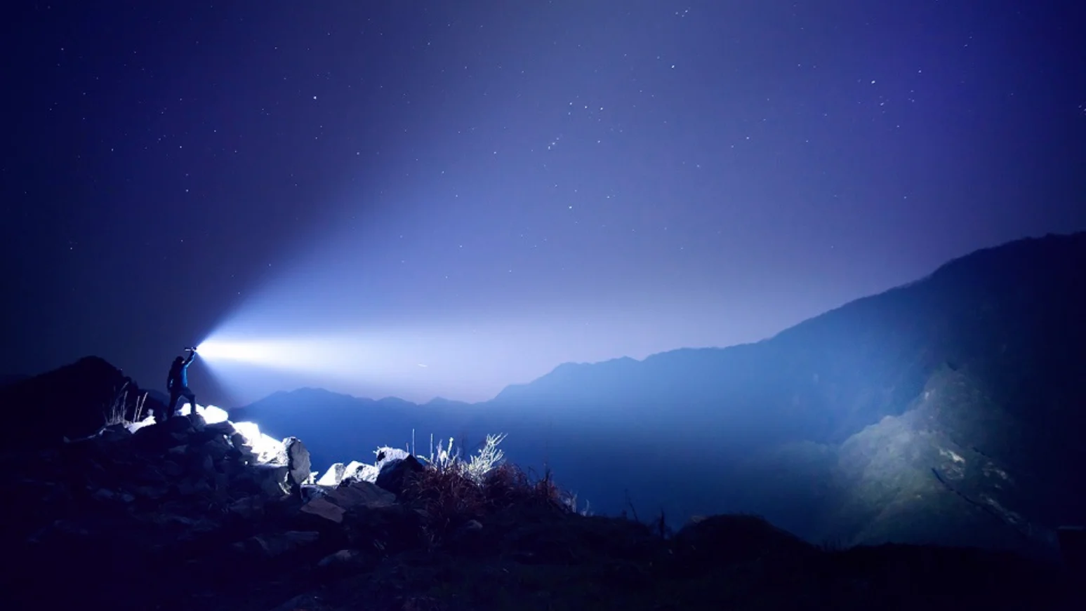 Person shines powerful flashlight on mountain peak at night, under starry sky.